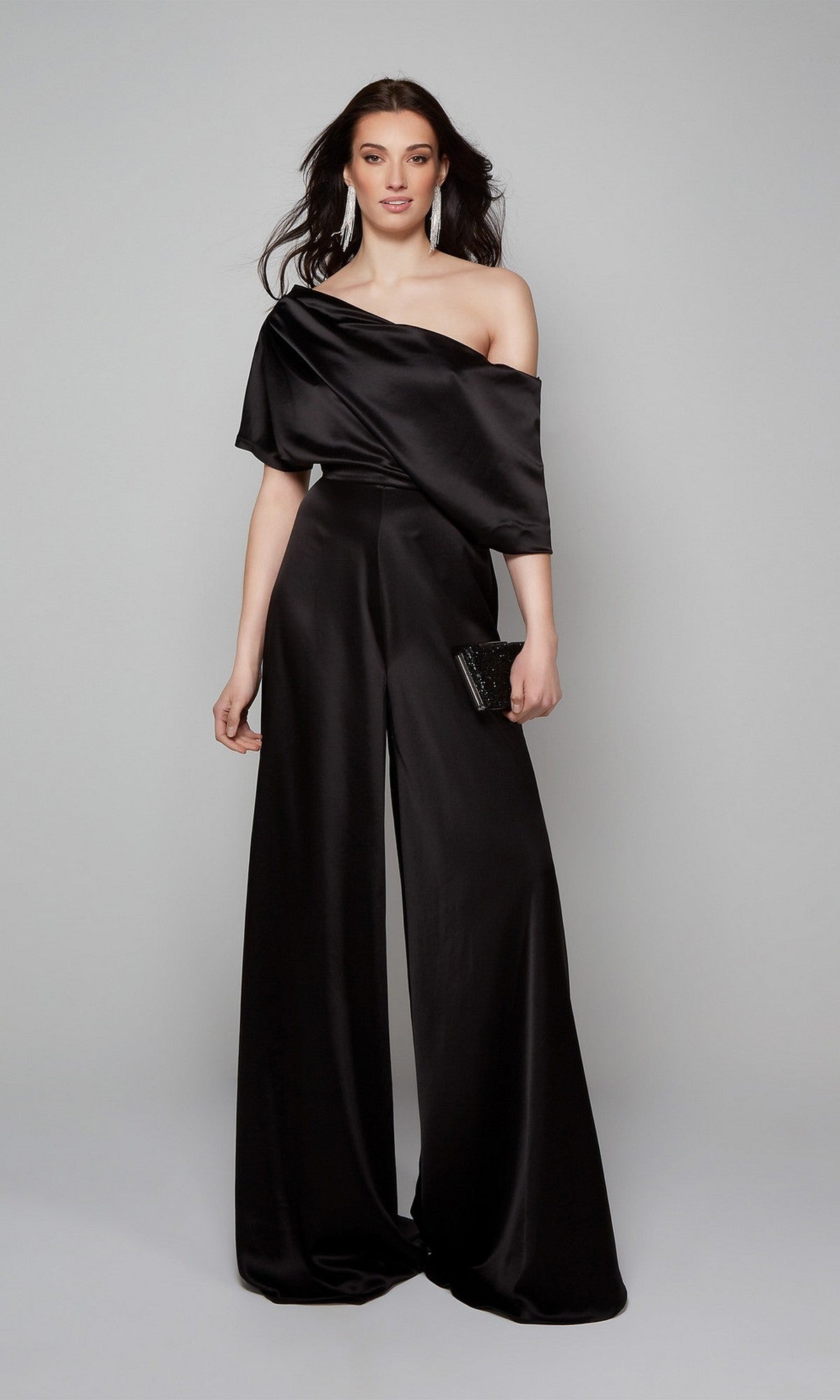 A woman wearing the Alyce 70019 Long Formal Jumpsuit with wide-leg pants holds a black clutch and stands against a plain gray background.